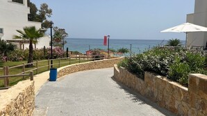 Un sendero pavimentado que conduce a una playa, con un muro de piedra a un lado y un edificio de fachada blanca al otro.