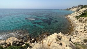 Paisaje costero con aguas turquesas cristalinas, costa rocosa y edificios a lo lejos.