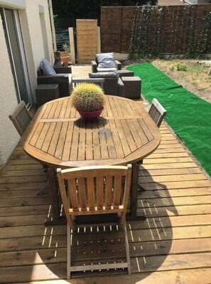 Eine Holzterrasse mit rundem Tisch und Stühlen, einem Blumentopf mit einem Kaktus und einem grünen Rasen im Hintergrund.