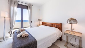 Un dormitori amb llit, una tauleta de nit, una làmpada, una finestra amb cortines i vista al mar.