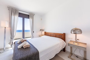 Een slaapkamer met een bed, nachtkastje, lamp, raam met gordijnen en uitzicht op zee.