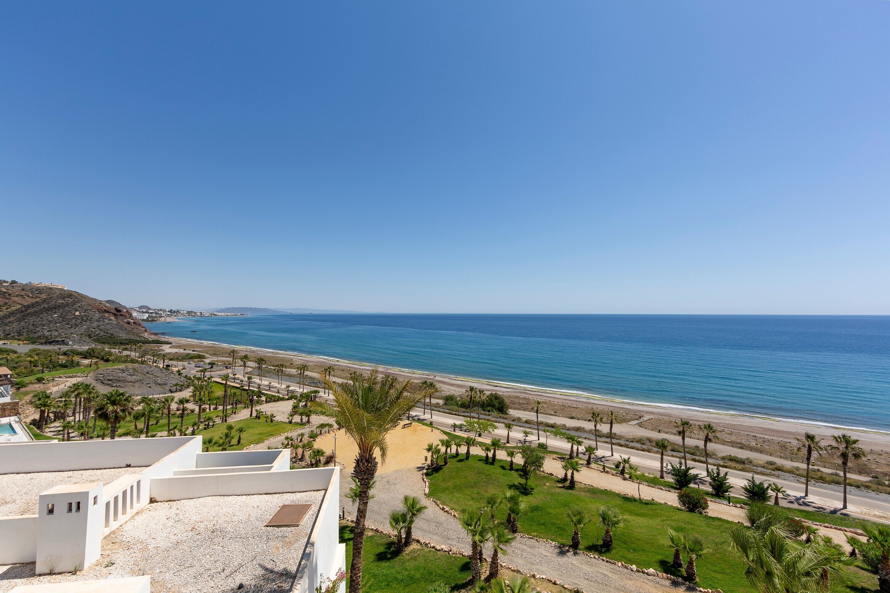 A coastal view with a beach, palm trees, and a white building with a pool.