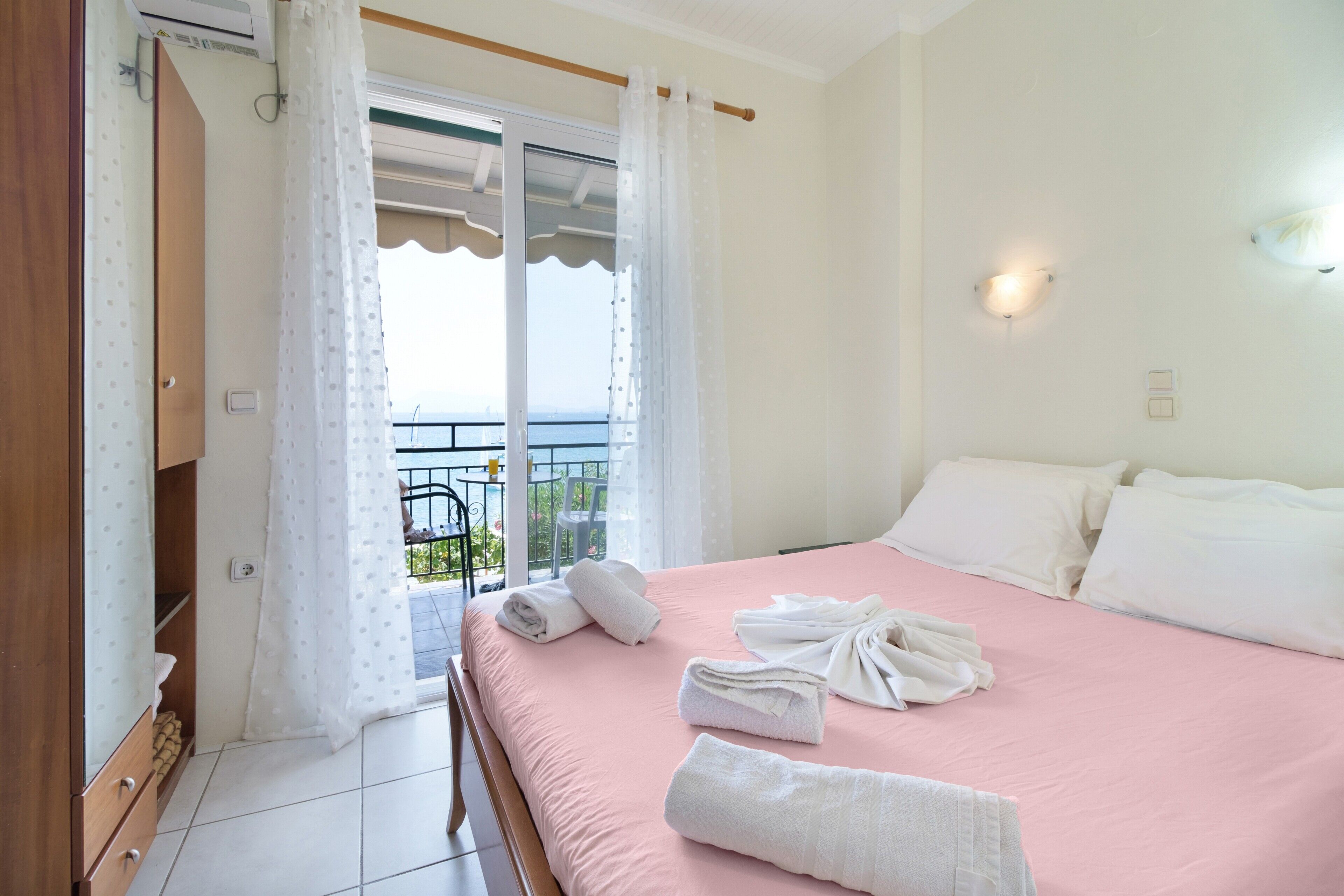 A bedroom with a pink bed, white towels, a balcony with a railing, and a view of the sea.