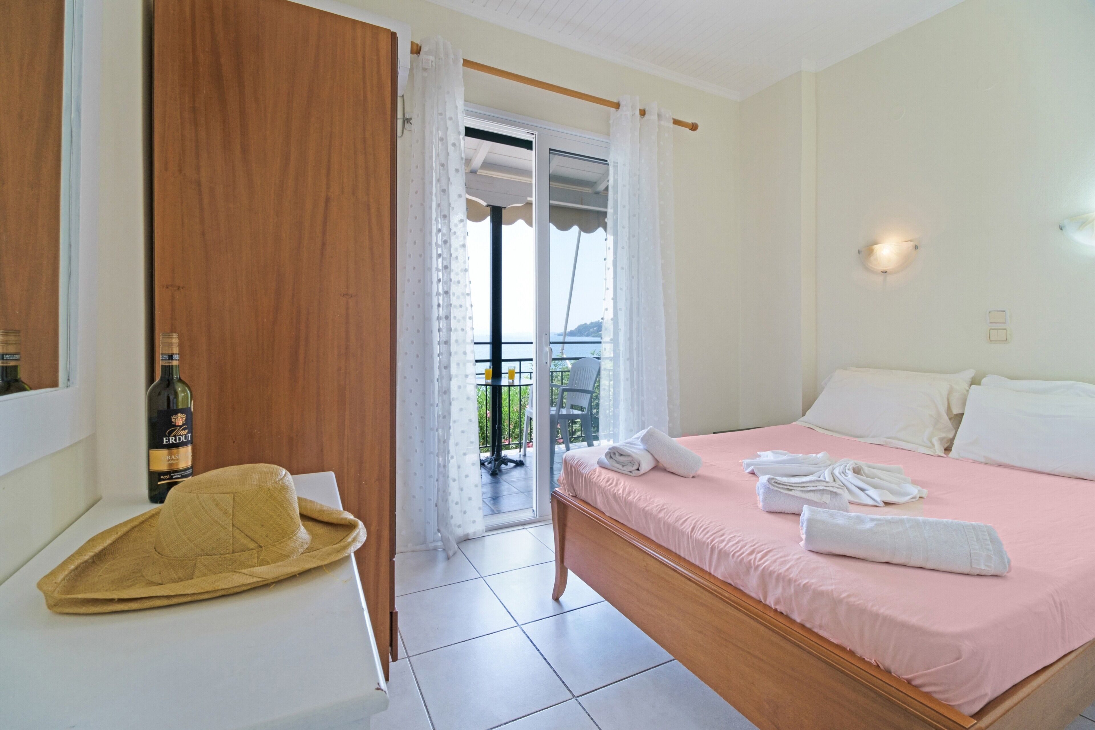 A bedroom with a bed, a wooden wardrobe, a hat, a bottle, and a view through a glass door.