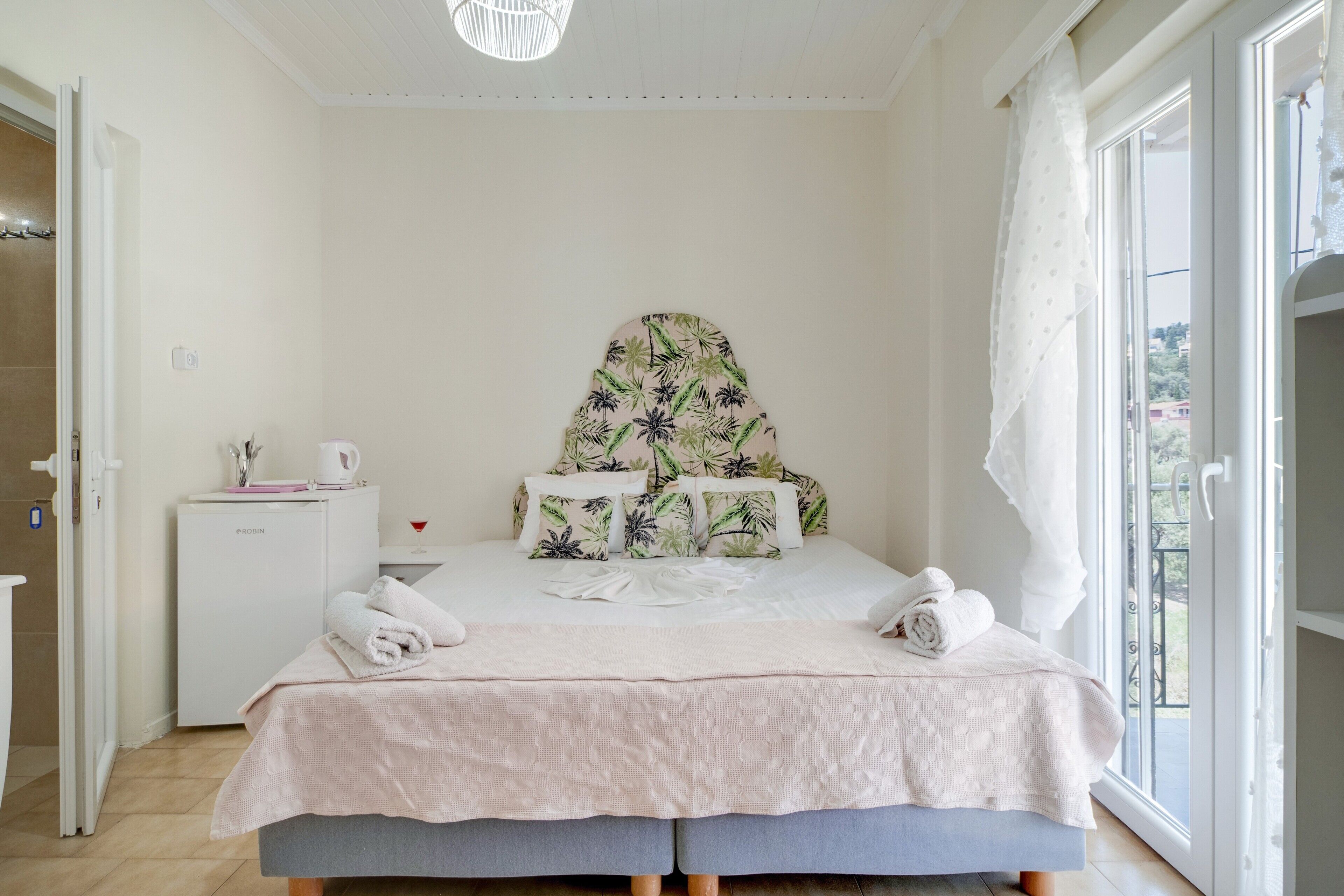 A bedroom with a bed, a small white bedside table, a window with a view, and a door.