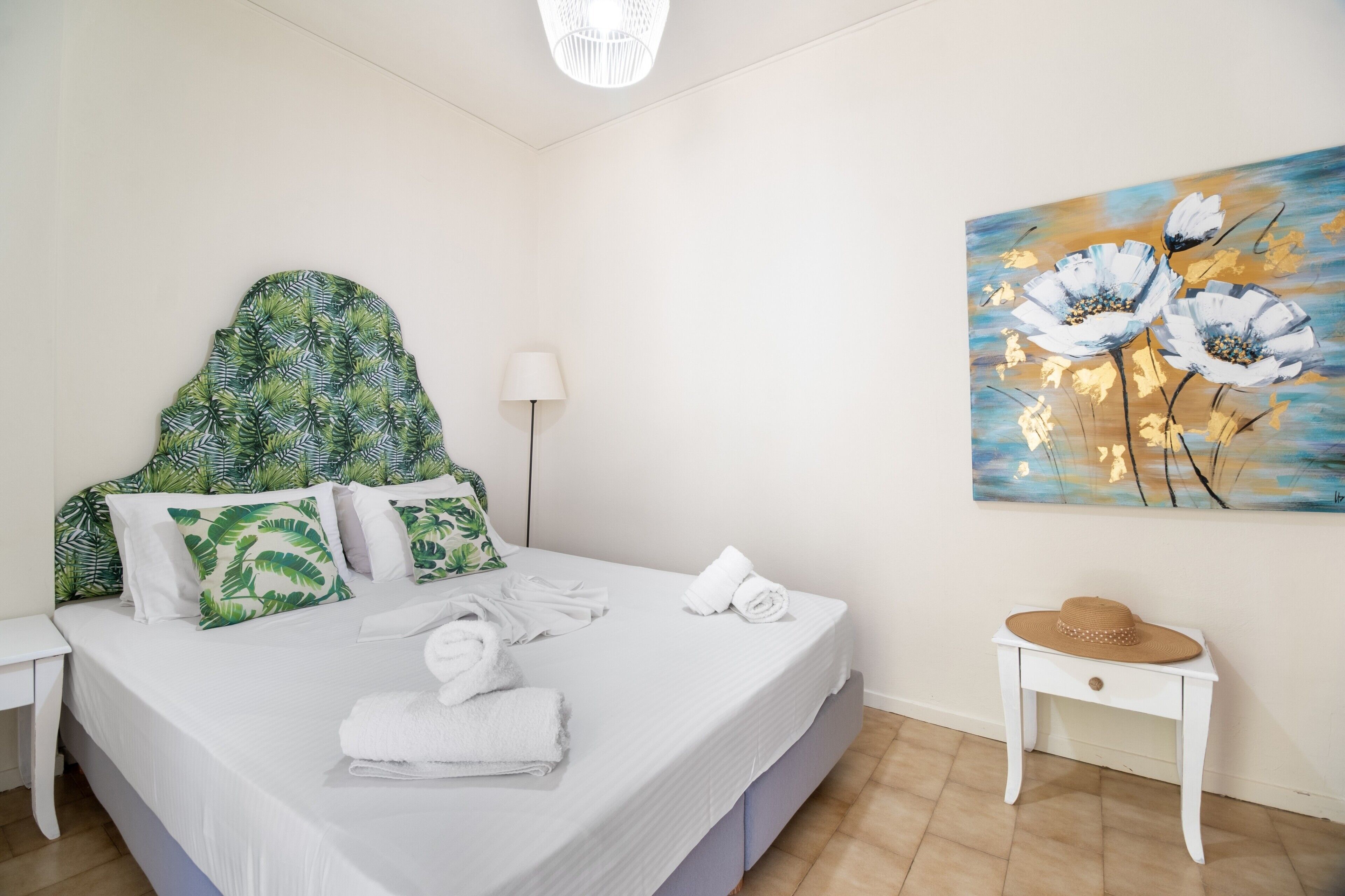 A neatly made bed with white linens, a green and white patterned headboard, and a painting of flowers on the wall.
