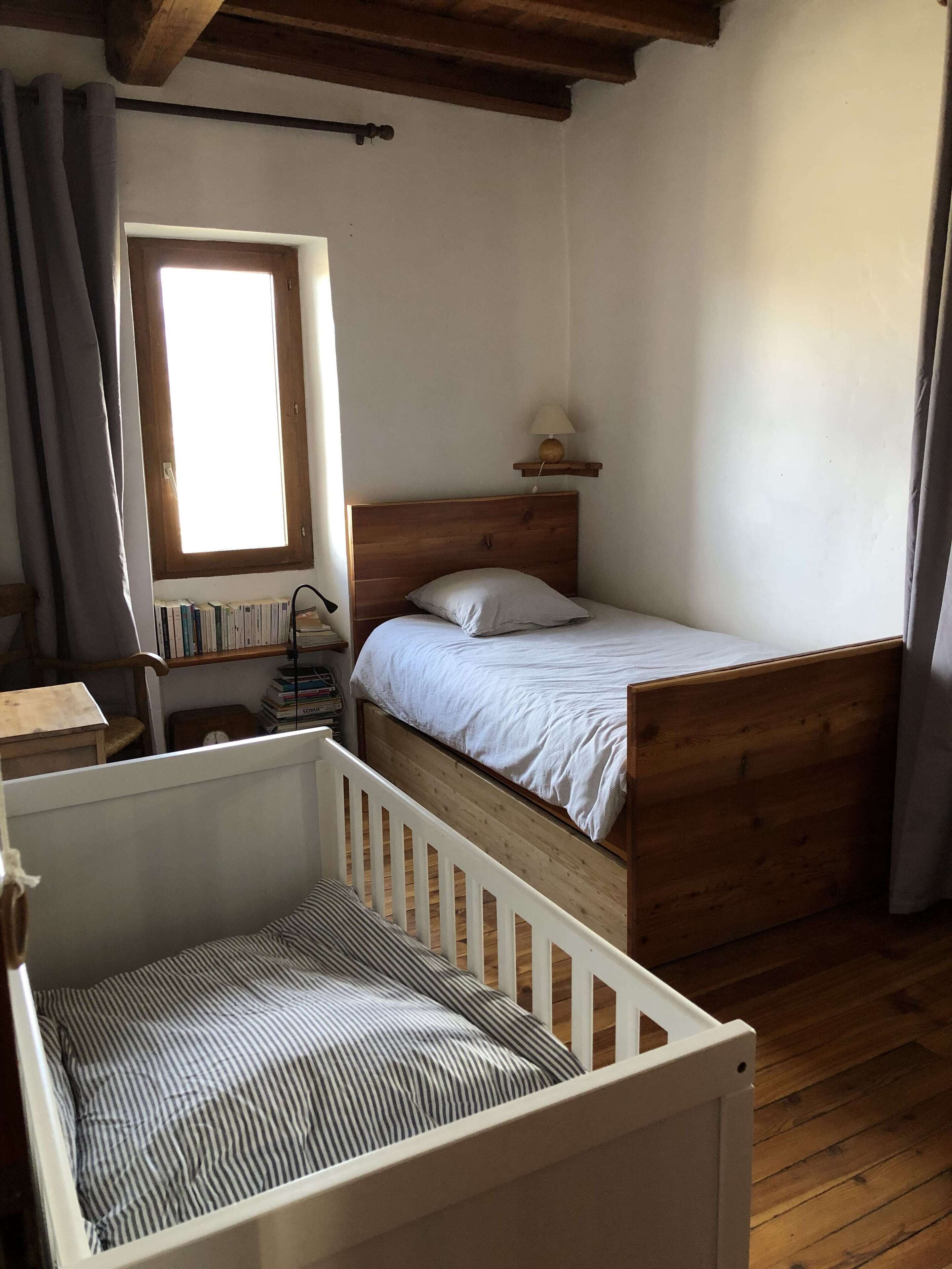 Ein Zimmer mit einem Holckrippe, einem Einzelbett, einem kleinen Tisch und einem Fenster mit Bücherregalen.