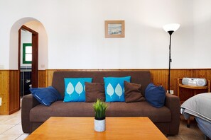 A living room with a brown sofa, blue and brown pillows, a wooden coffee table with a plant, a framed picture on the wall, and a floor lamp.