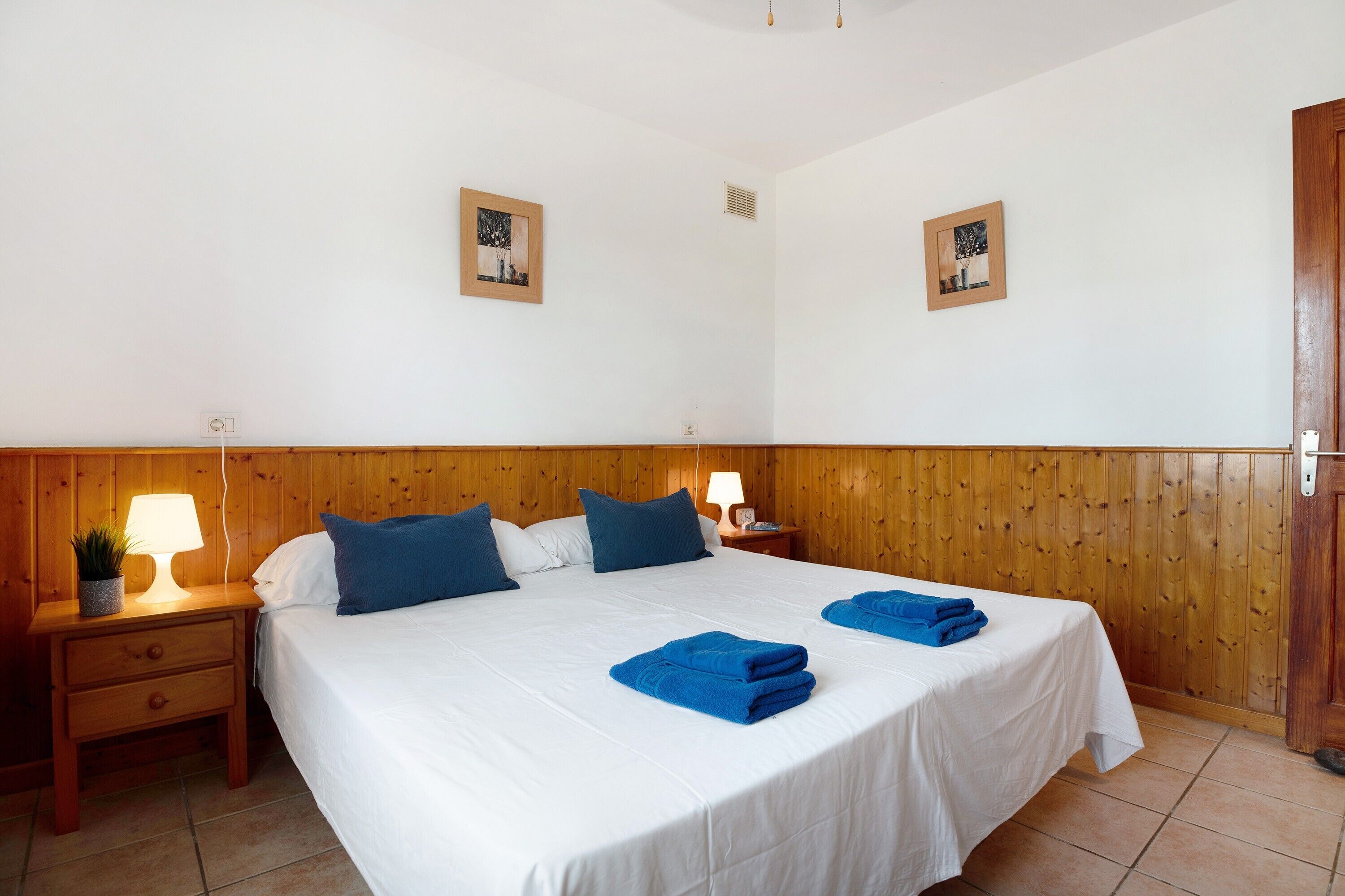 Ein Schlafzimmer mit einem hölzernen Kopfende, einem Bett mit weißer Bettwäsche und blauen Kissen, einem Nachttisch mit Lampe und zwei gerahmten Bildern an der Wand.