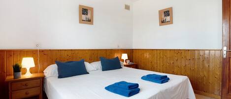 Ein Schlafzimmer mit einem hölzernen Kopfende, einem Bett mit weißer Bettwäsche und blauen Kissen, einem Nachttisch mit Lampe und zwei gerahmten Bildern an der Wand.