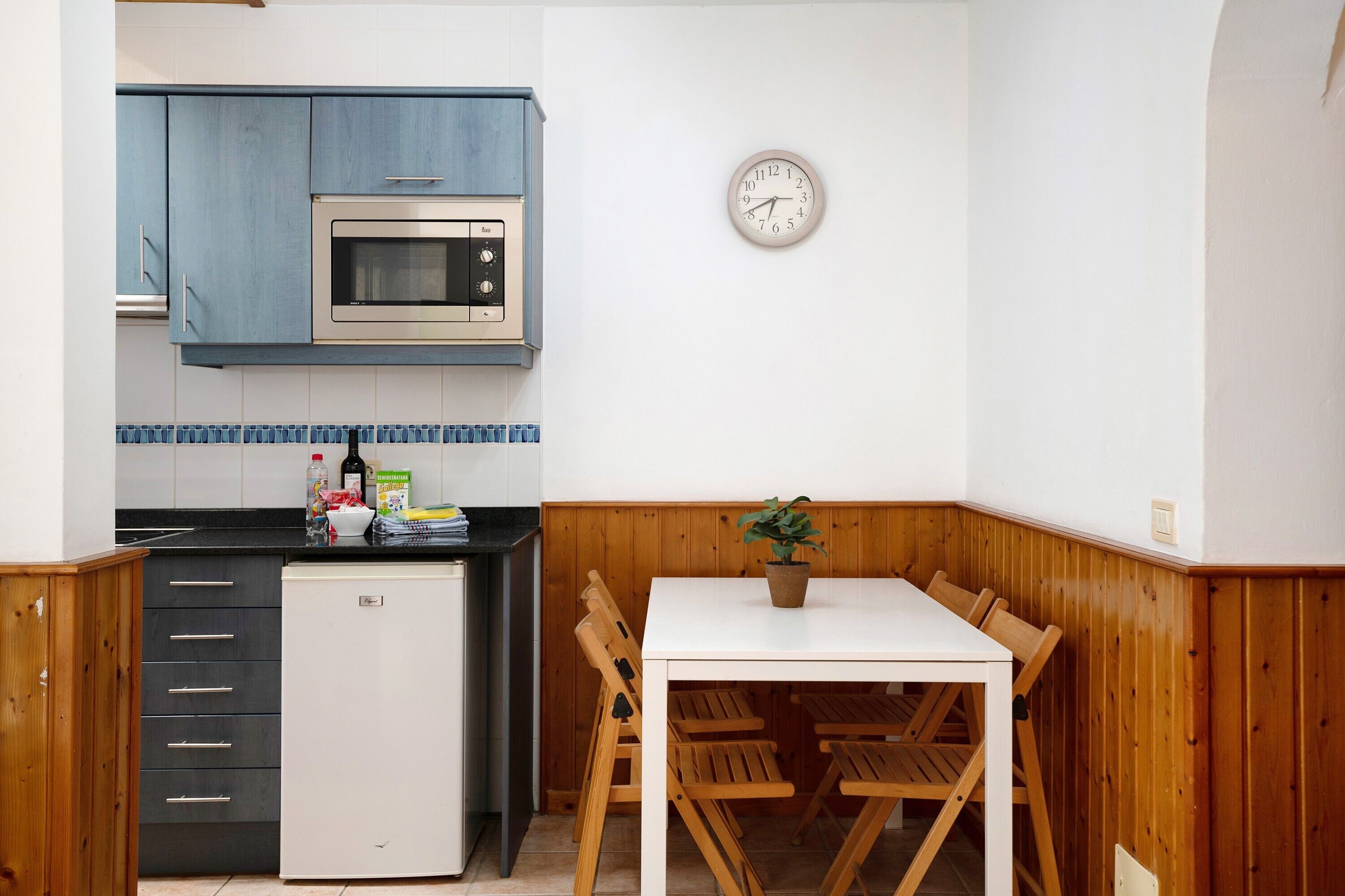 A kitchen with a microwave, refrigerator, wooden cabinets, a clock, and a dining table with chairs.