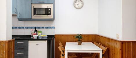 A kitchen with a microwave, refrigerator, wooden cabinets, a clock, and a dining table with chairs.