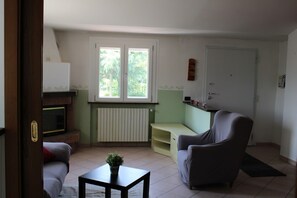 A living room with a fireplace, a sofa, a chair, a small table with a plant, and a window with a view of greenery.