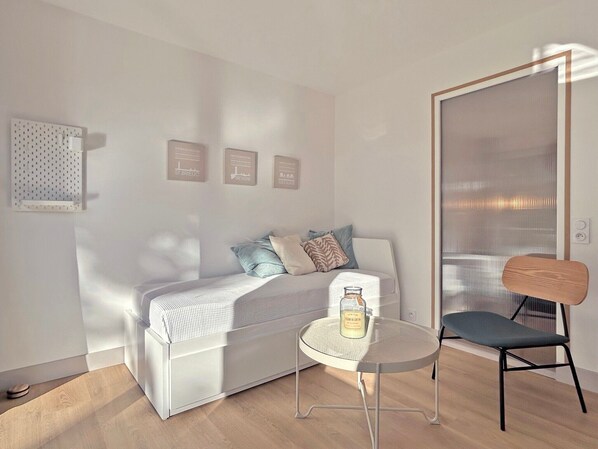 Ein modernes Zimmer mit einem weißen Sofa, einem kleinen runden Tisch mit einer Flasche, einem Sessel und einer Wandleuchte.