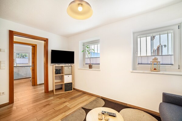 A modern living room with a flat-screen TV, a wooden entertainment unit, a small window with a plant, and a view of the outside. - Apartment "Unterortsgut" with Private Garden & Wi-Fi (Dorf Tirol)