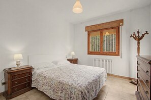 A bedroom with a bed, a dresser, a nightstand, a lamp, a window with curtains, and a radiator.