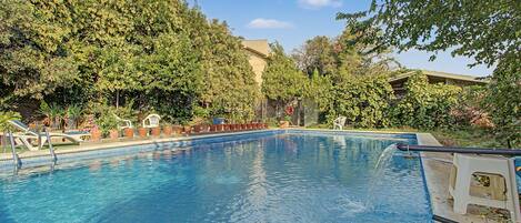 A swimming pool with clear blue water, surrounded by lush green trees and a building in the background.