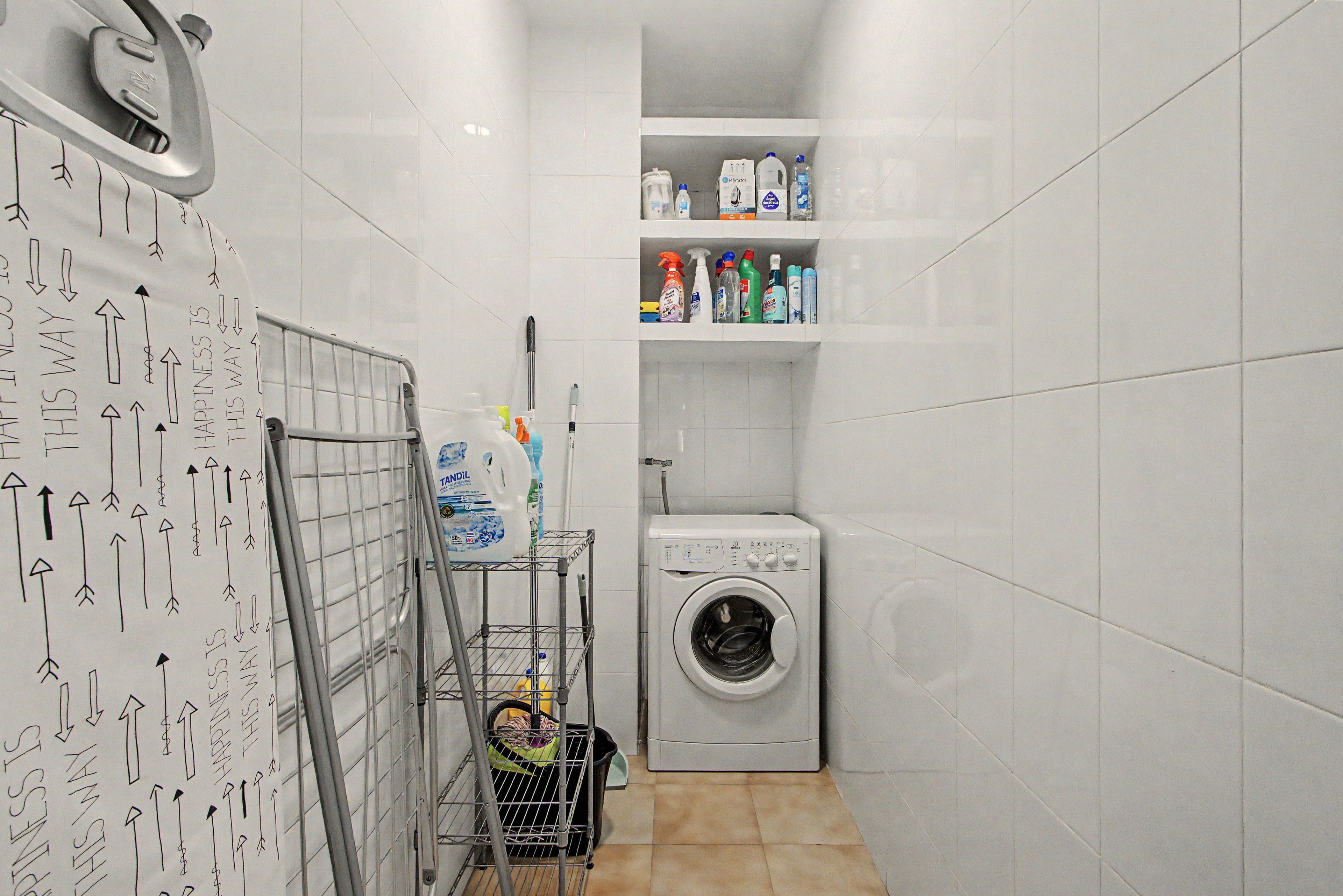Een wasruimte met een wasmachine, planken met schoonmaakproducten en een metalen rek met opgevouwen kleding.