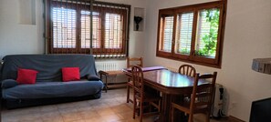 A living room with a sofa, dining table, chairs, and two windows with blinds. - Rural House 'Marina' with Private Terrace, Private Garden and Wi-Fi (Lleida)