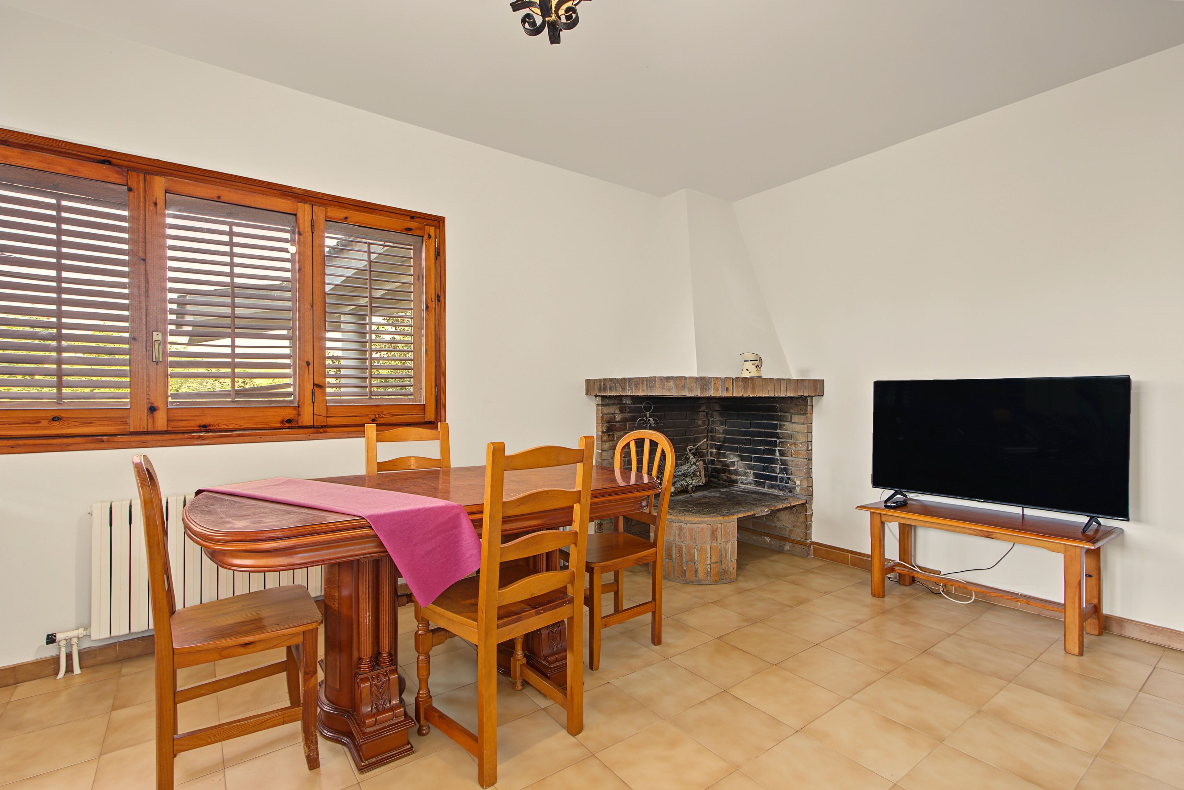 Un coin repas avec une table en bois, des chaises et une nappe rose. Une cheminée se trouve à droite, avec un téléviseur posé sur un meuble.