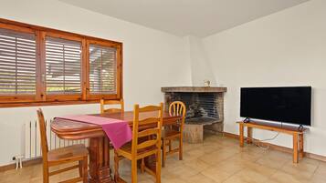 Un coin repas avec une table en bois, des chaises et une nappe rose. Une cheminée se trouve à droite, avec un téléviseur posé sur un meuble.