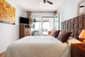 A bedroom with a bed, a television, a ceiling fan, a large window, and a view of the outside.