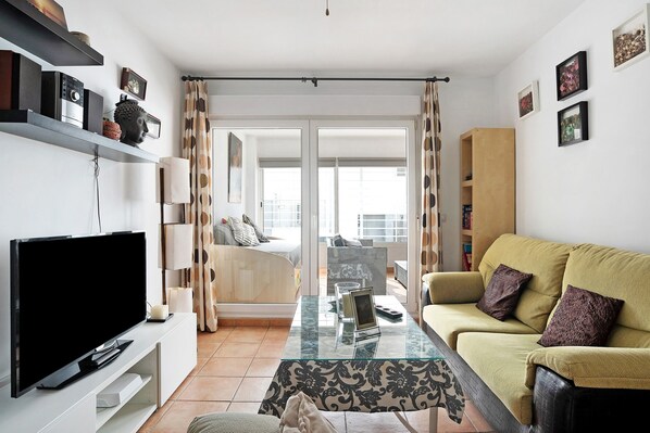 A living room with a flat-screen TV, sofa, glass coffee table, and a view through a sliding door to an outdoor balcony.
