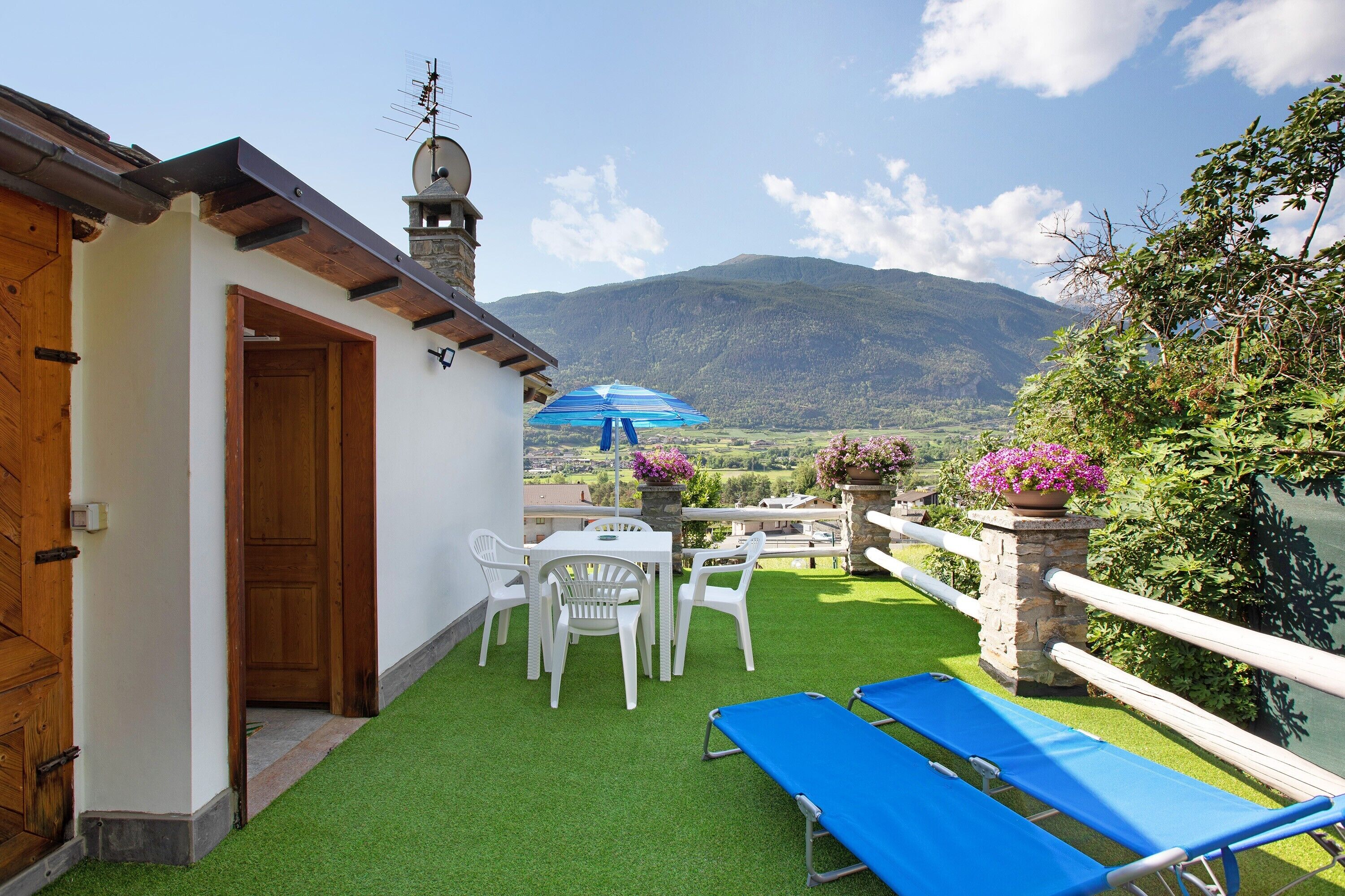 A terrace with artificial grass, a table with chairs, a blue lounge chair, and a view of mountains.