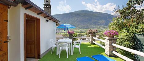 A terrace with artificial grass, a table with chairs, a blue lounge chair, and a view of mountains.