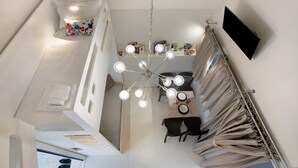 Una habitación de hotel moderna con una decoración en tonos blancos, un candelabro, un televisor de pantalla plana y un área de comedor con una mesa y sillas.