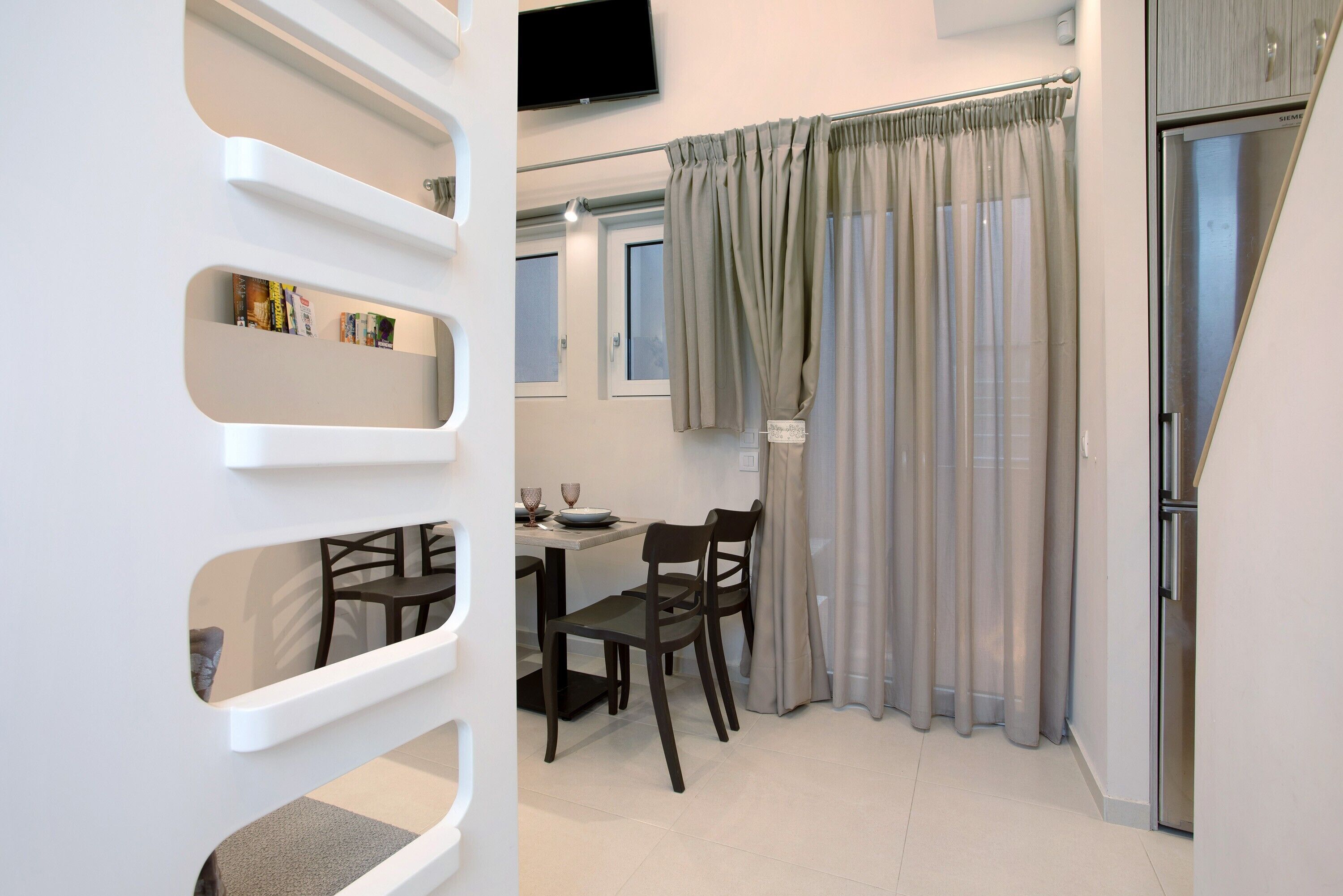 A modern kitchen with a dining area, a built-in bookshelf, and a flat-screen TV mounted on the wall.