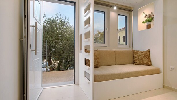 Una habitación con puerta de vidrio corrediza que da a un balcón, un sofá con cojines estampados, un estante pequeño de pared con una planta y una ventana con vistas a un edificio y árboles.