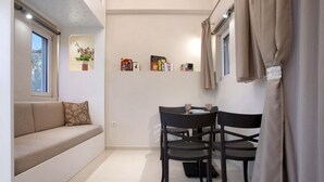 Un espai habitable reduït amb un sofà, una taula de dinar amb cadires, una petita prestatgeria i una finestra amb cortines.