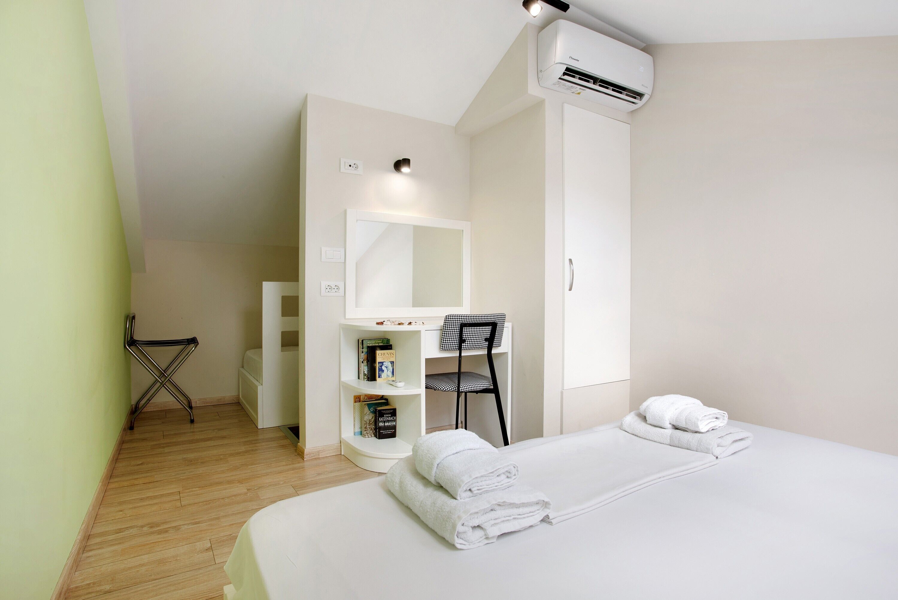 A hotel room with a bed, a desk with a chair, a bookshelf, and an air conditioning unit.