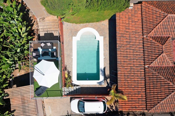 Eine Vogelperspektive zeigt ein Anwesen mit Schwimmbad, einer Terrasse mit Gartenmöbeln und einem in der Nähe stehenden Auto.