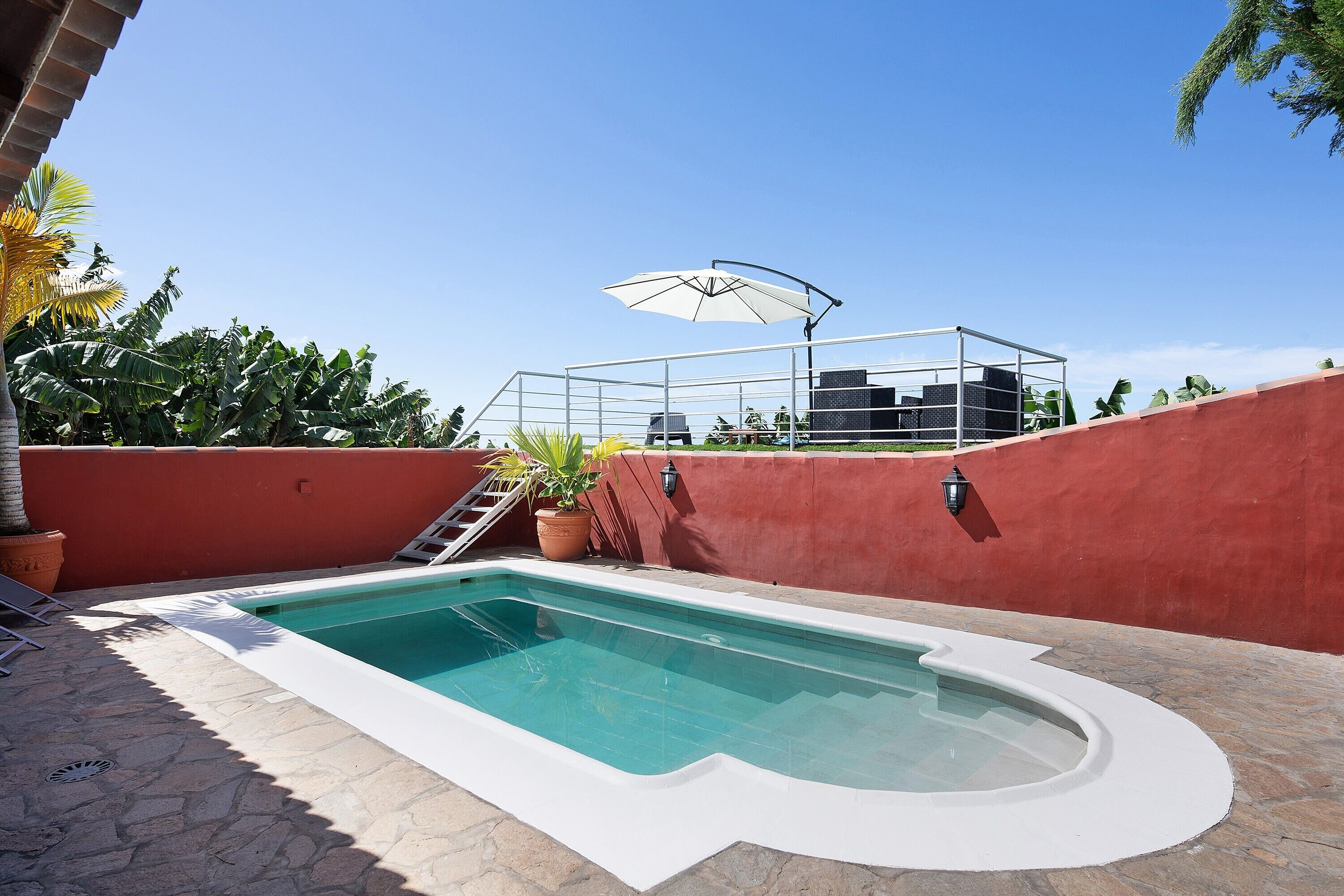 A swimming pool with a stone deck, a red wall, and a metal structure with an umbrella.