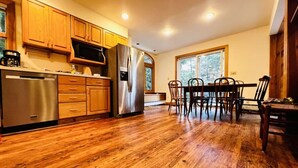 A kitchen with wooden cabinets, stainless steel appliances, and a dining area with chairs and a table. - Stratton Mountain Home - Hot Tub, Fire Pit & Game Loft (Jamaica)