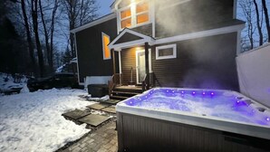 A hot tub in front of a house with a snow-covered yard and a wooden deck. - Stratton Mountain Home - Hot Tub, Fire Pit & Game Loft (Jamaica)