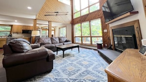 A spacious living room with a large sofa, a coffee table, and a fireplace. There is a television mounted on the wall and a kitchen area visible in the background. - Stratton Mountain Home - Hot Tub, Fire Pit & Game Loft (Jamaica)