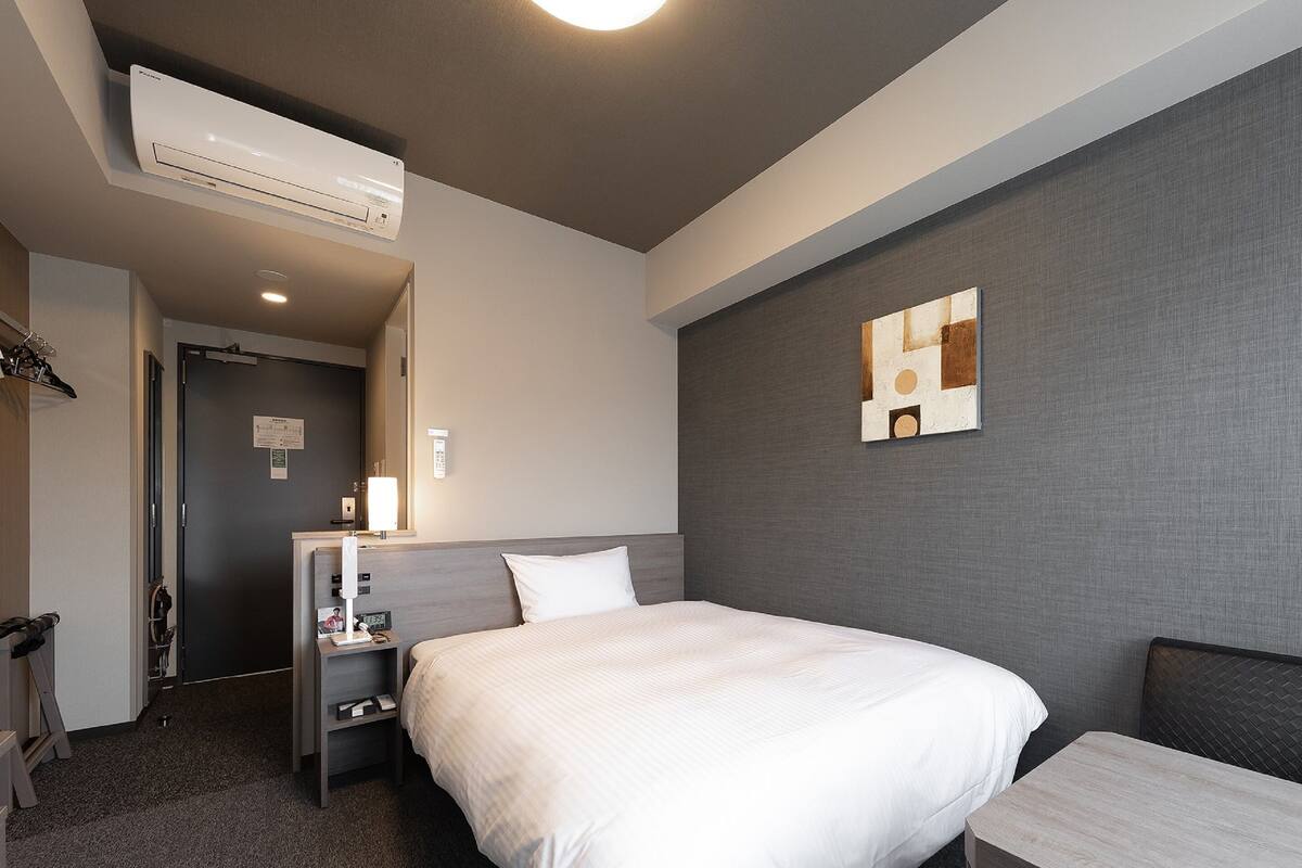 A hotel room with a bed, bedside table, lamp, and an air conditioning unit.