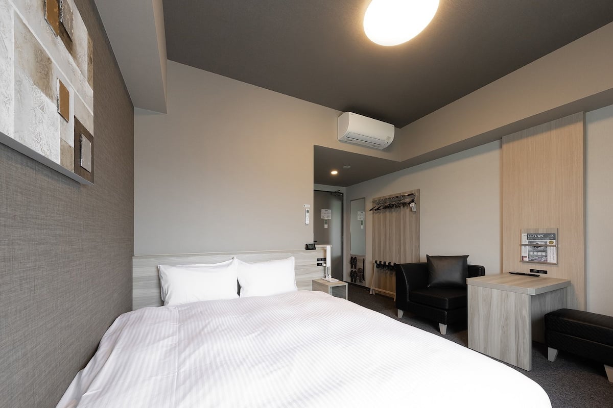 A hotel room with a large bed, a desk, a chair, and an air conditioning unit.