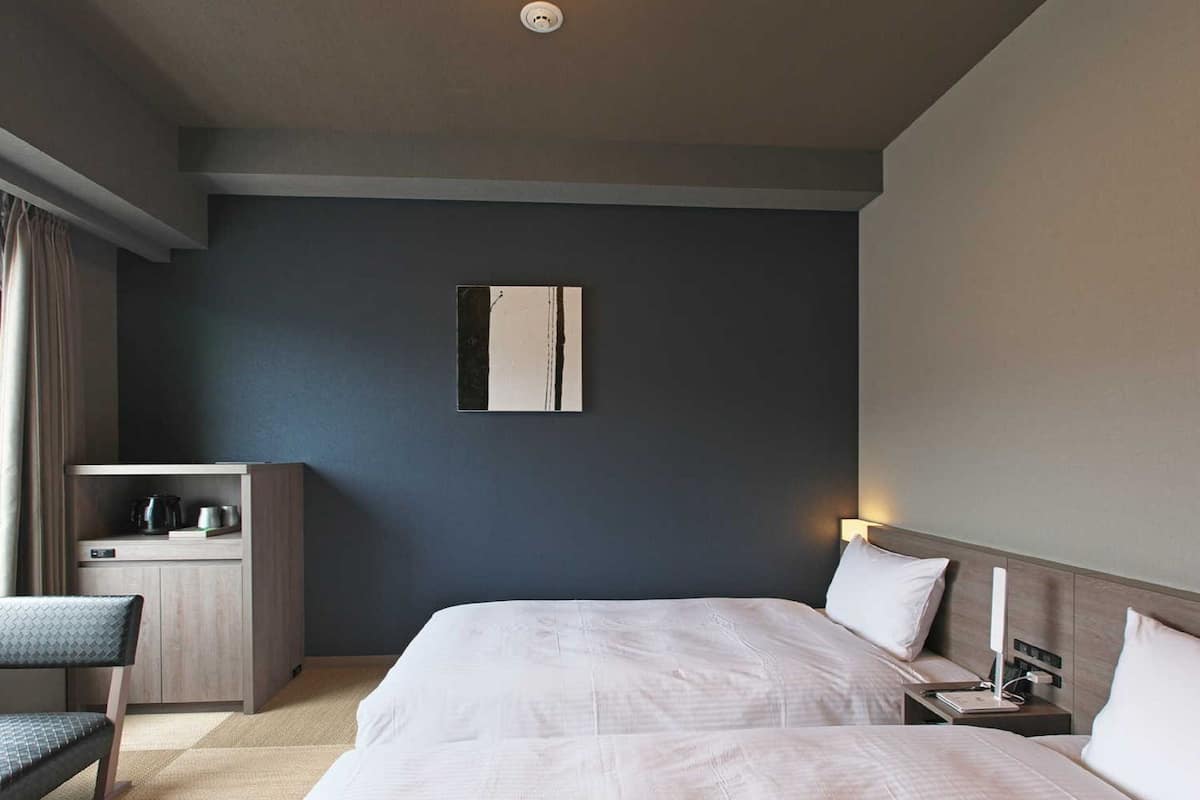 A hotel room with two beds, a desk, a chair, a bedside table, and a wall-mounted artwork.