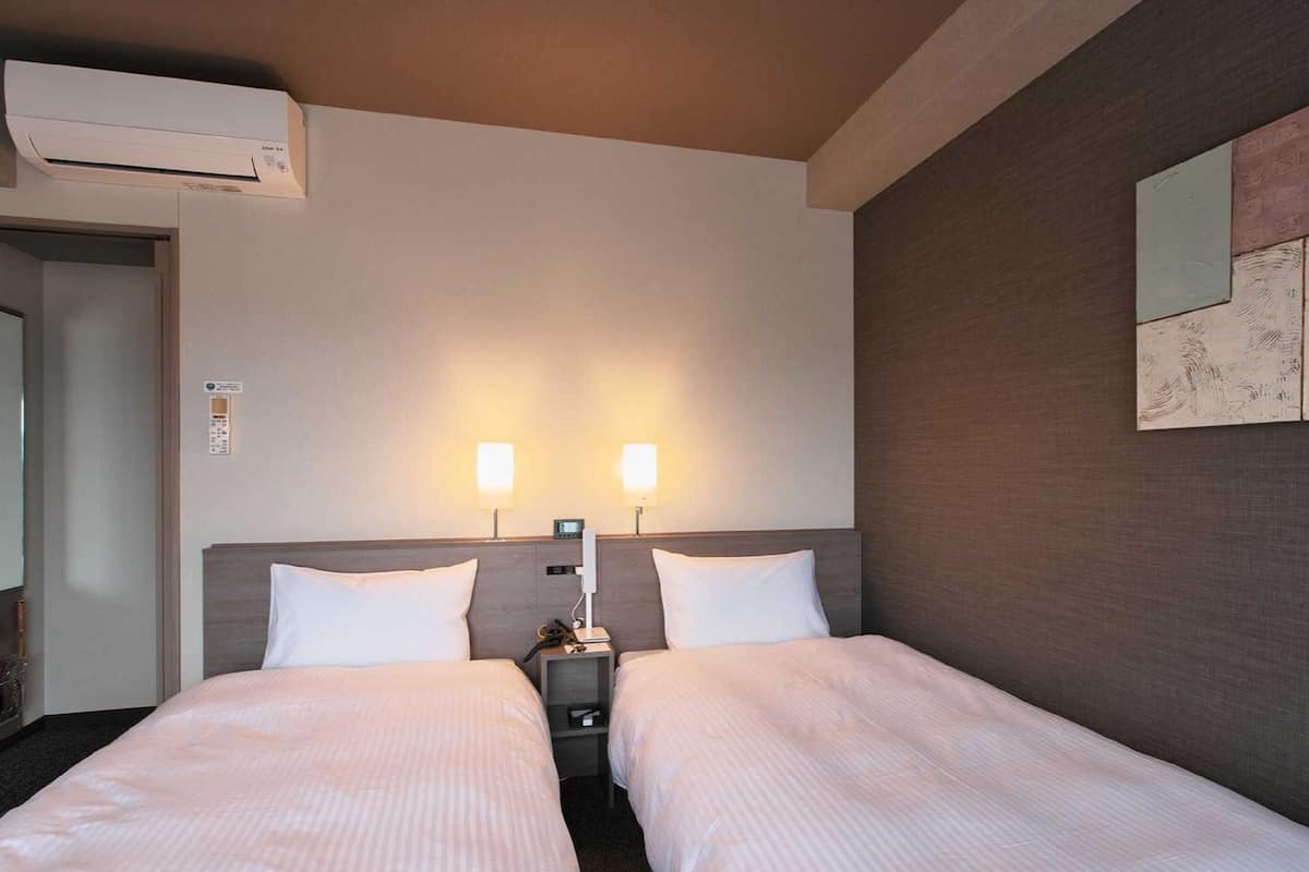 A hotel room with two beds, a wall-mounted air conditioner, bedside lamps, and a small table with a phone.