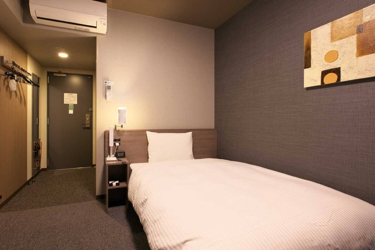 A hotel room with a bed, a nightstand with a lamp, an air conditioner, and a wall with a geometric painting.