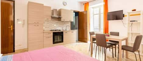 A compact hotel room with a kitchenette, a dining area, and a bed with a pink blanket.