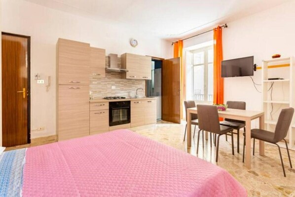 Ein kompaktes Hotelzimmer mit Küchenzeile, einer Essecke und einem Bett mit rosa Decke.