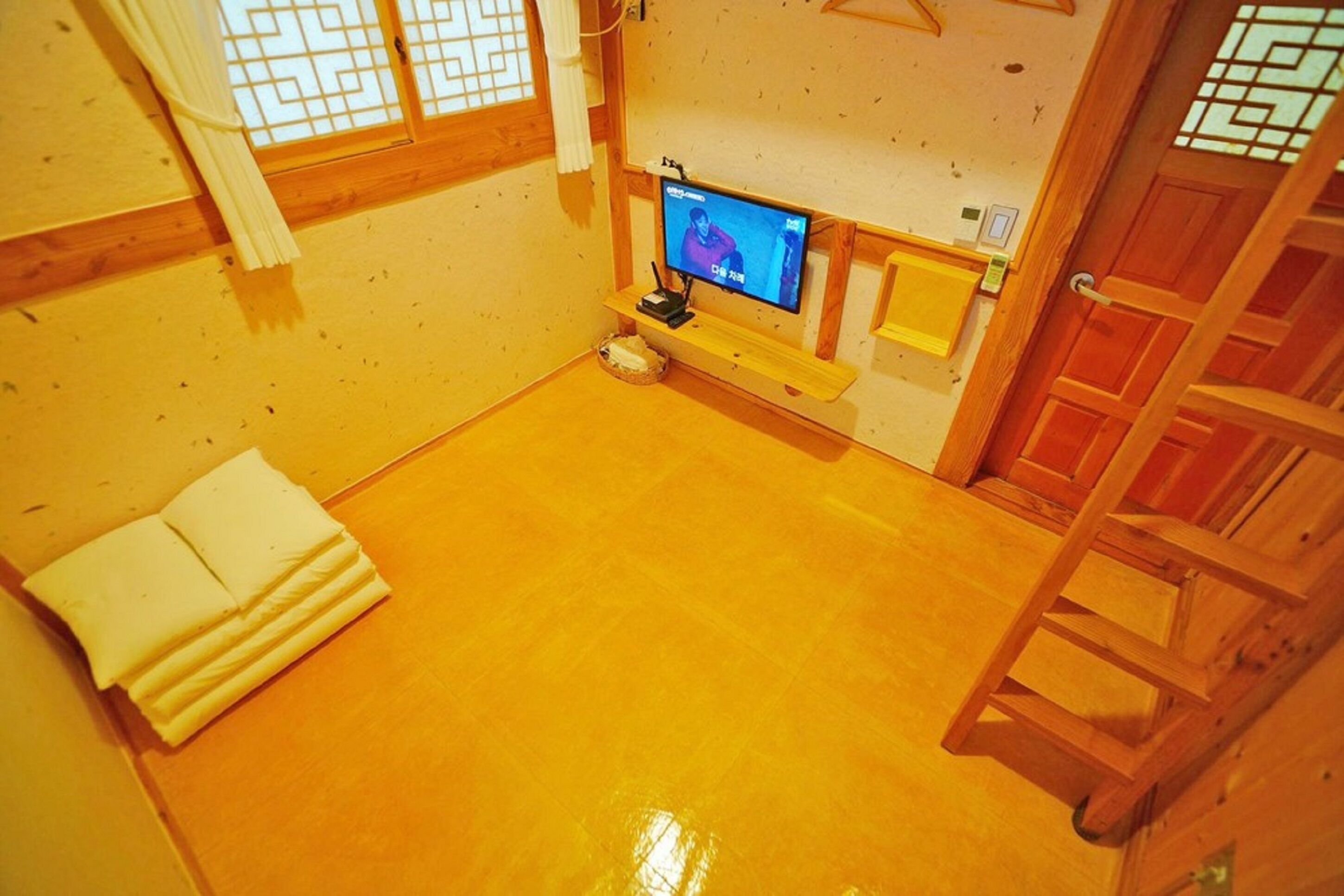 A room with a wooden floor, a television on a stand, a white cushion, and a wooden ladder.