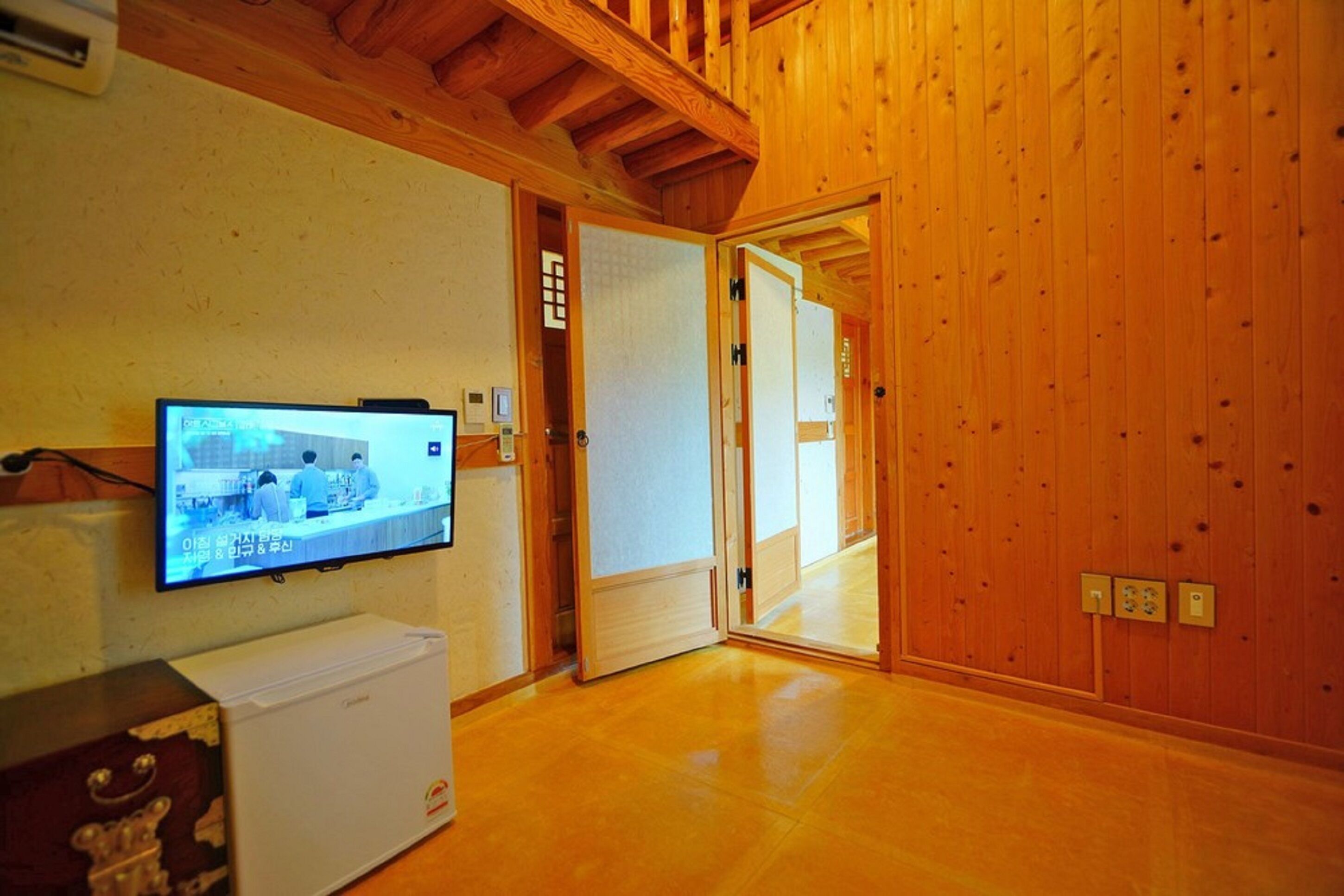 A room with wooden walls and a wooden ceiling, a television mounted on the wall, and a white refrigerator.