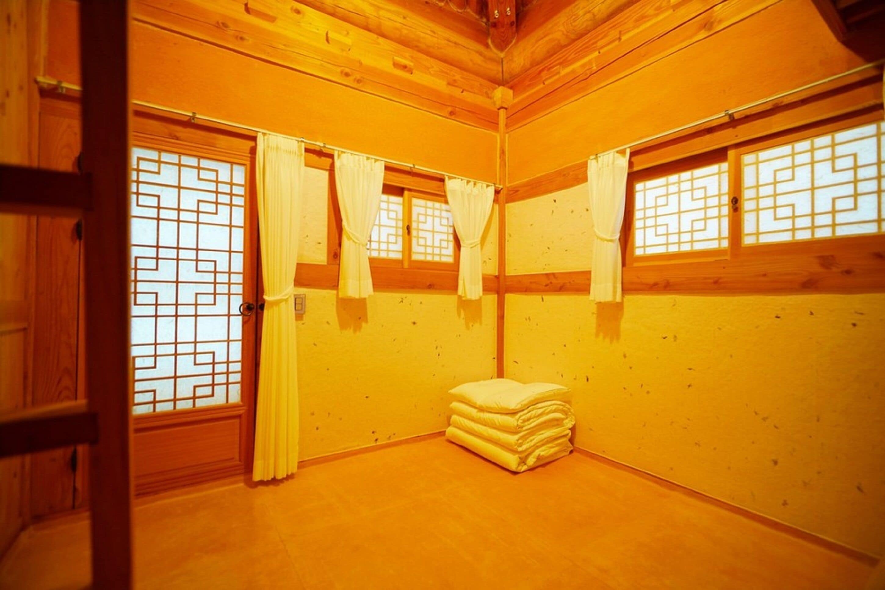 A room with wooden walls and a wooden floor, featuring a window with a geometric pattern, a door, and a stack of pillows.