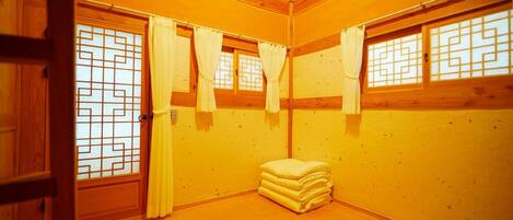 A room with wooden walls and a wooden floor, featuring a window with a geometric pattern, a door, and a stack of pillows.